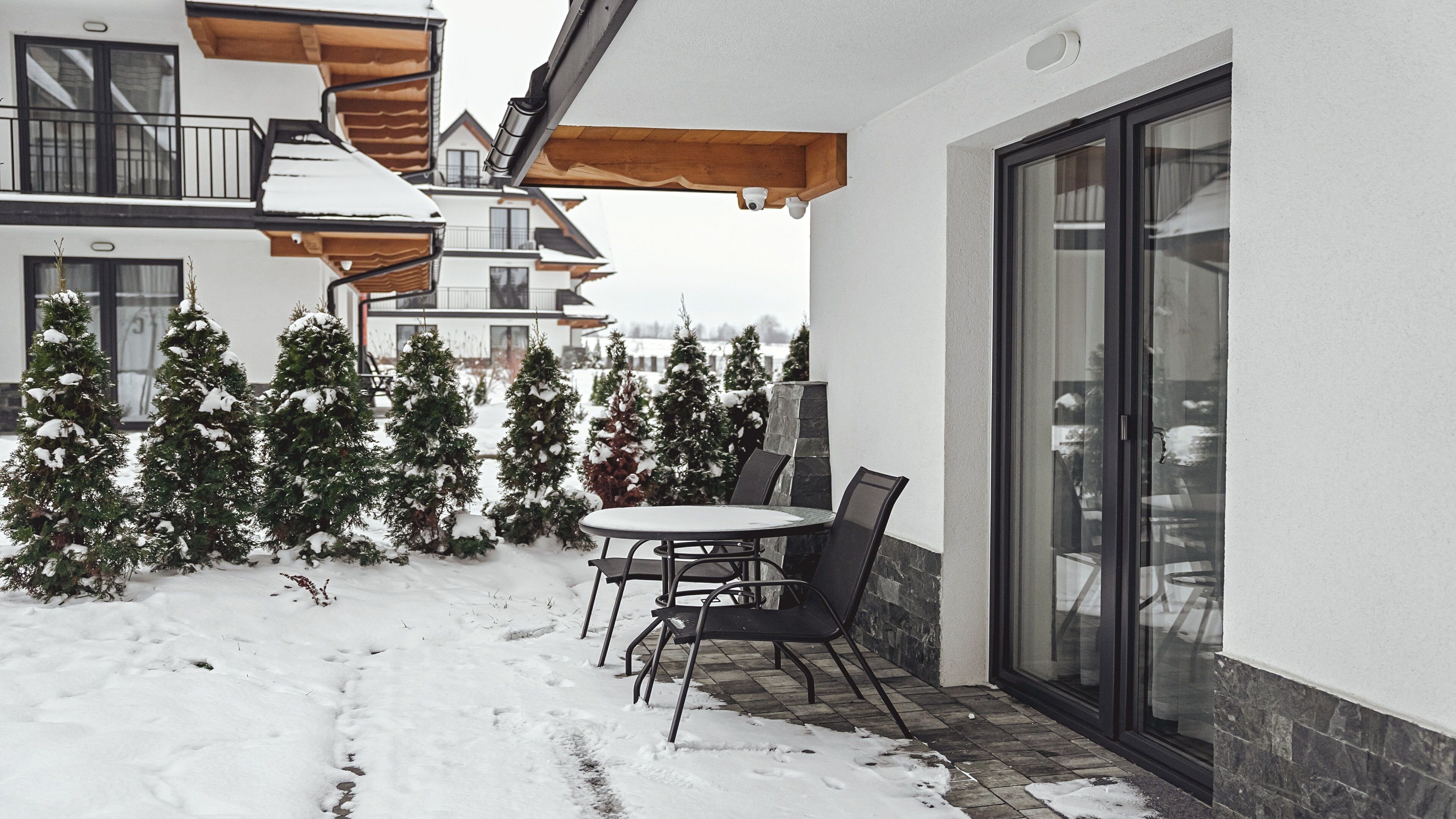 A snow-covered patio with a table and chairs, surrounded by snow-laden evergreen trees.