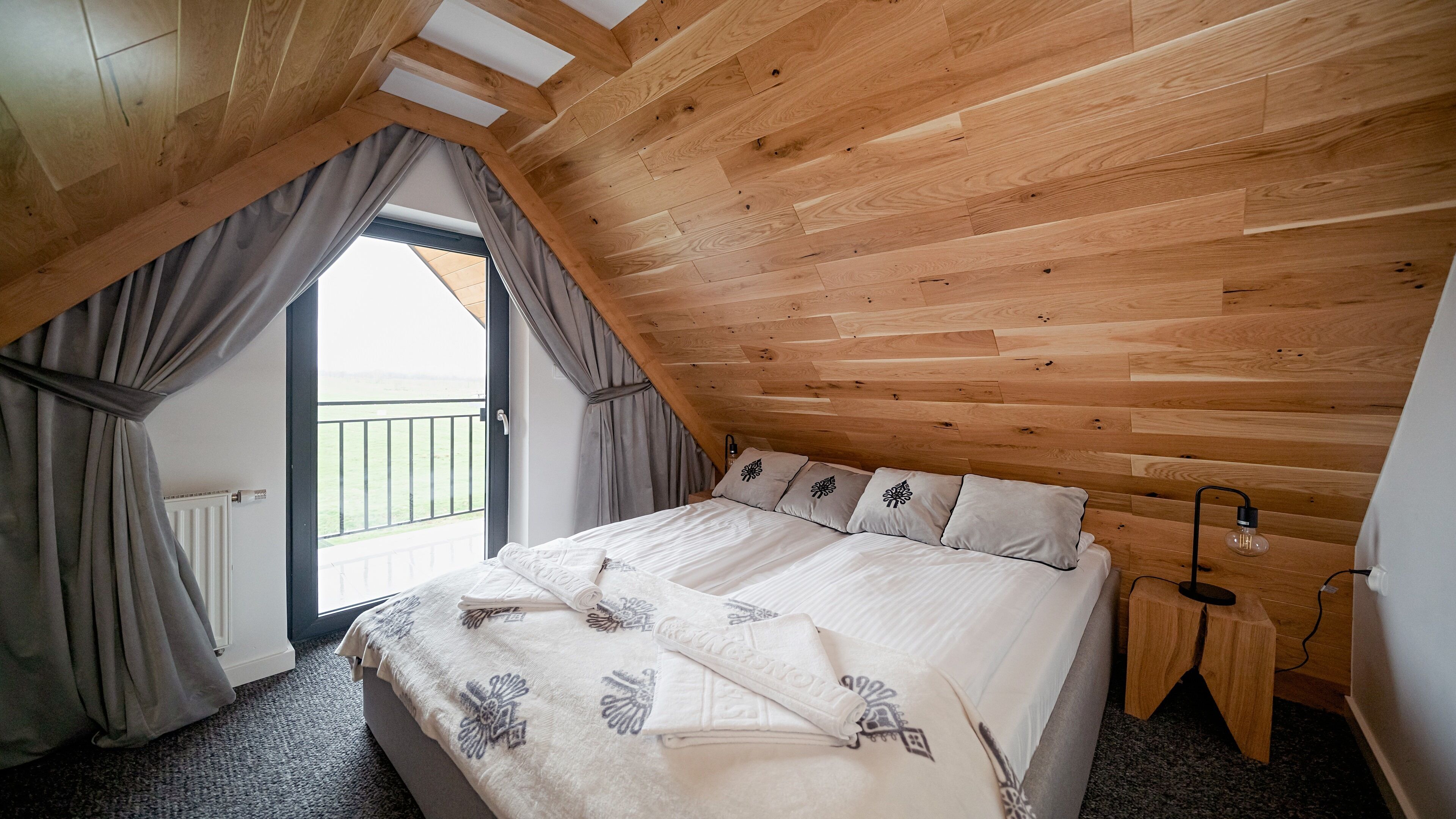 A bedroom with a sloped ceiling, a bed with a white bedspread and pillows, a small wooden stool, and a view of a balcony.