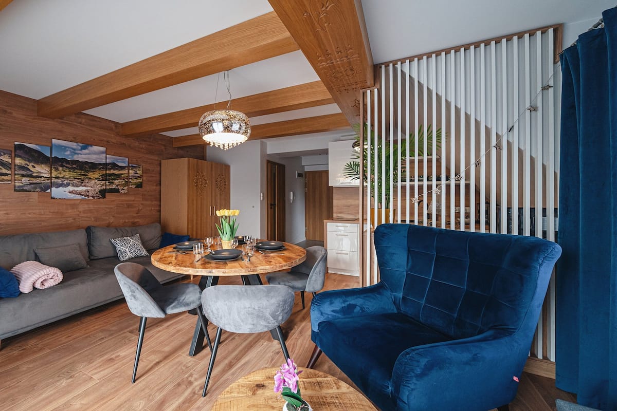 Un salon moderne avec un plafond en bois, une table à manger ronde dressée pour quatre personnes, un fauteuil bleu, un canapé gris et un escalier avec une rampe blanche.