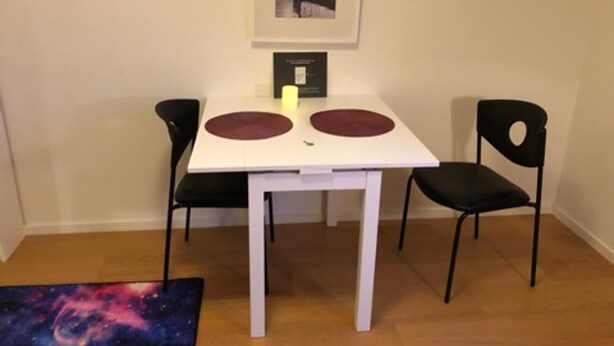 Un pequeño comedor con una mesa blanca, dos sillas negras y un cuadro en la pared.
