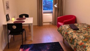 Una habitación pequeña y bien iluminada con una mesa de comedor, dos sillas, un sillón rojo, una cama con colcha estampada de flores y una alfombra con diseño galáctico.