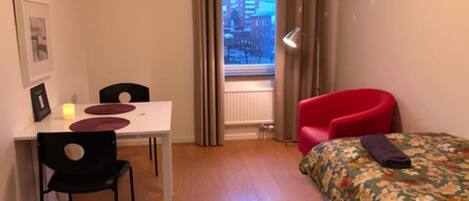 A small, well-lit room with a dining table, two chairs, a red armchair, a bed with a floral bedspread, and a galaxy-patterned rug.