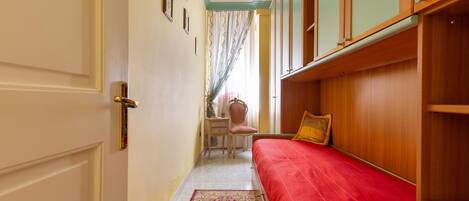 A narrow hallway leading to a bedroom with a red bed, wooden wardrobe, and a chair with a cushion.