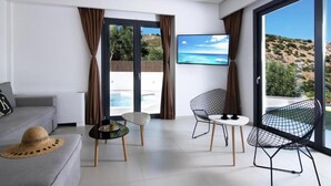 A modern living room with a grey sofa, a small round table, two wireframe chairs, and a large TV screen displaying a beach scene. - Deluxe Villa Konstantinos (Malevizi)