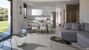 A modern living room with a grey sofa, a dining area with a table and chairs, and a kitchen in the background. - Deluxe Villa Konstantinos (Malevizi)