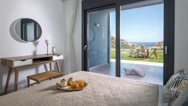 A bedroom with a bed, a bedside table, a mirror, and a view of a balcony with a table and chairs. - Deluxe Villa Konstantinos (Malevizi)