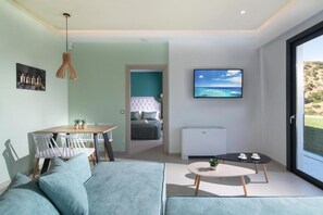 A modern living room with a sofa, a dining table with chairs, a TV mounted on the wall, and a view of the outdoors through a large window.
