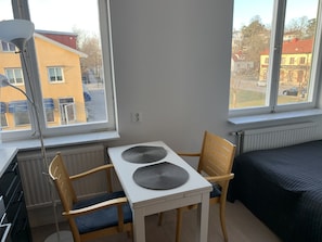 Et lille rum med et bord, to stole, en radiator og et vindue med udsigt til bygninger og parkerede biler.