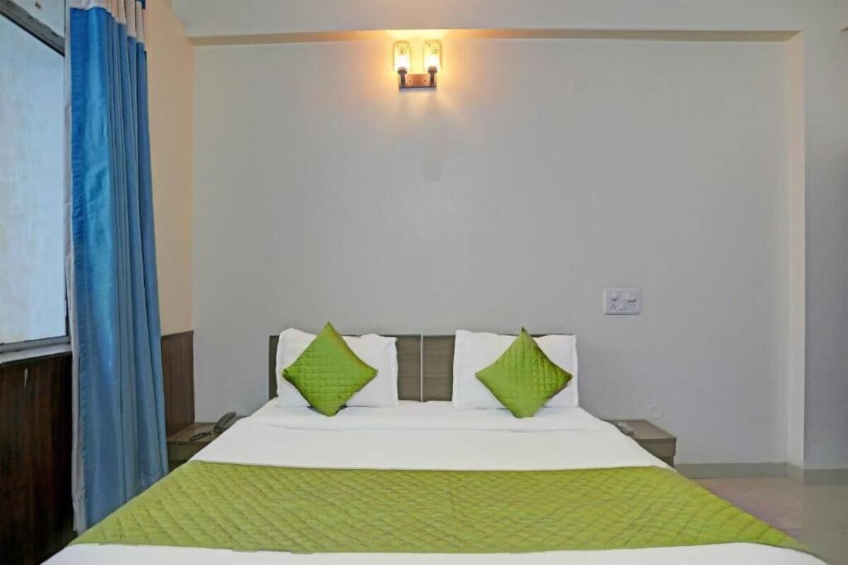 A bedroom with a bed, green pillows, a quilted green bedspread, a nightstand, a wall-mounted light fixture, and a blue curtain.