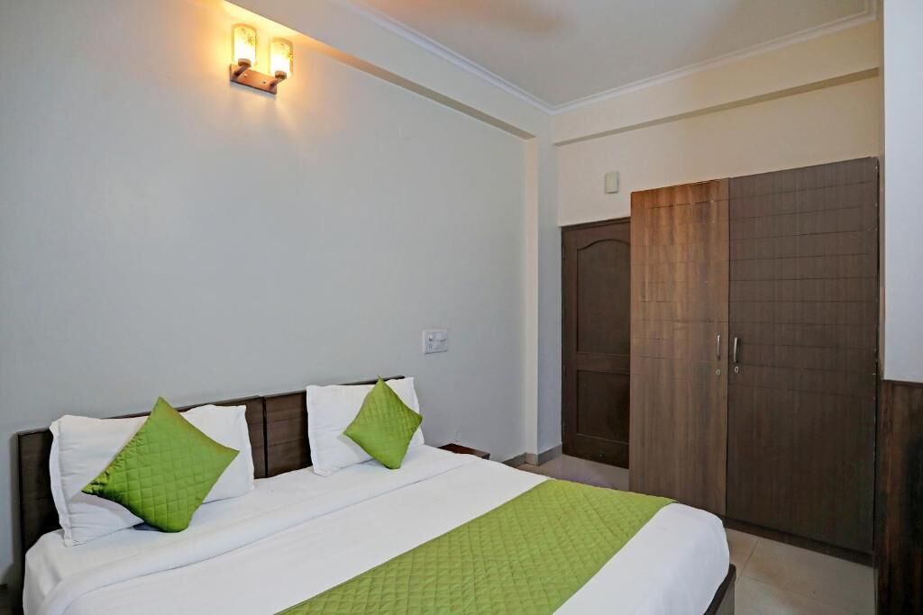 A hotel room with a bed, green pillows, a nightstand, a wardrobe, and a wall-mounted light fixture.