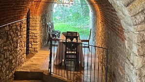 A stone-walled tunnel leading to an outdoor area with a table and chairs, a chandelier, and a view of greenery. - Il Castello di Bubbio (Bubbio)