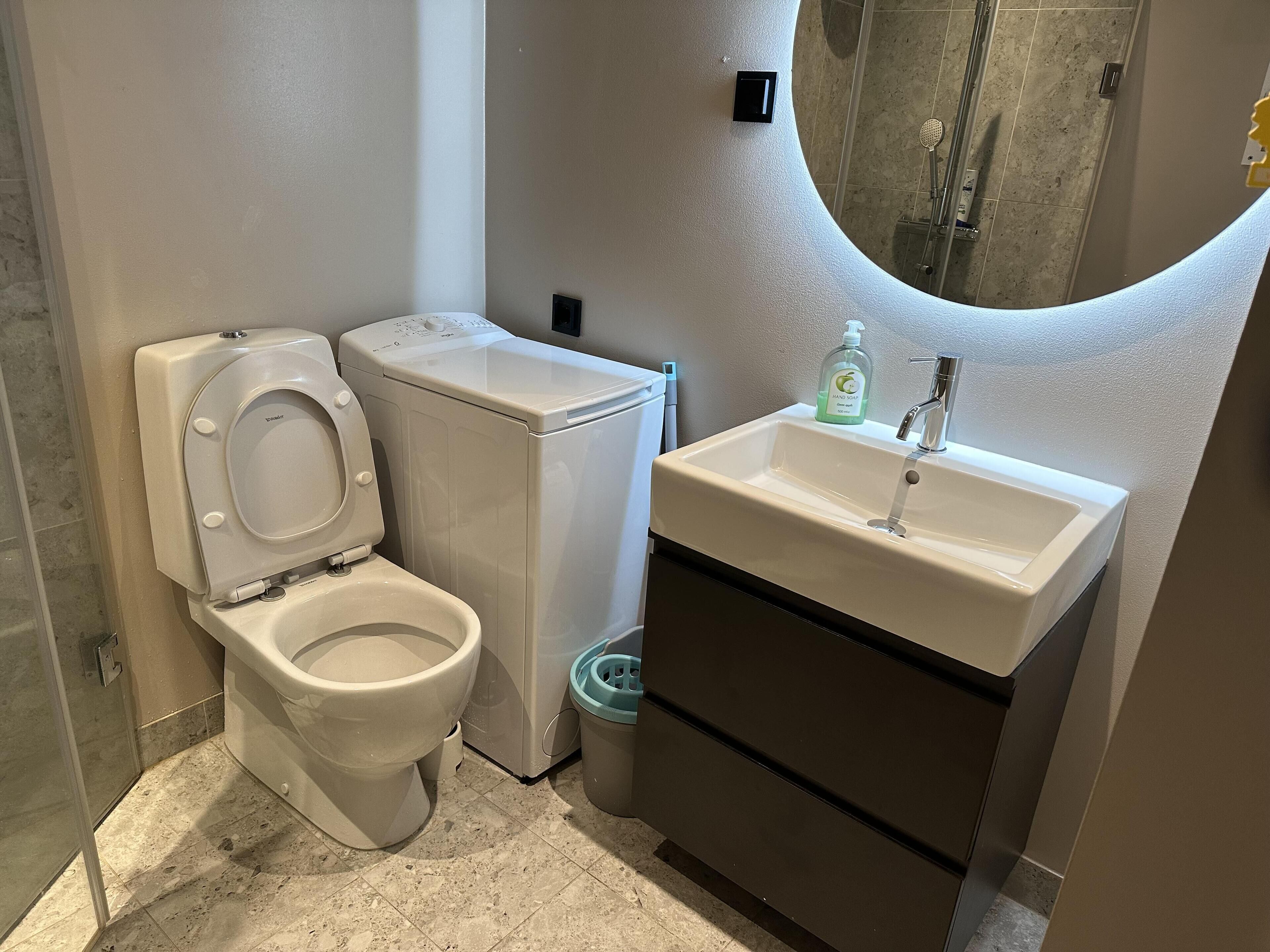 A compact bathroom with a toilet, washing machine, sink, and a mirror.