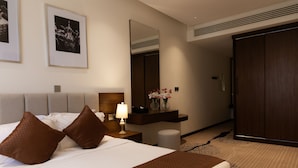 A hotel room with a large bed, a nightstand with a lamp, a mirror, a dresser, and a door leading to another room. - Dream Residence (Al Khobar)