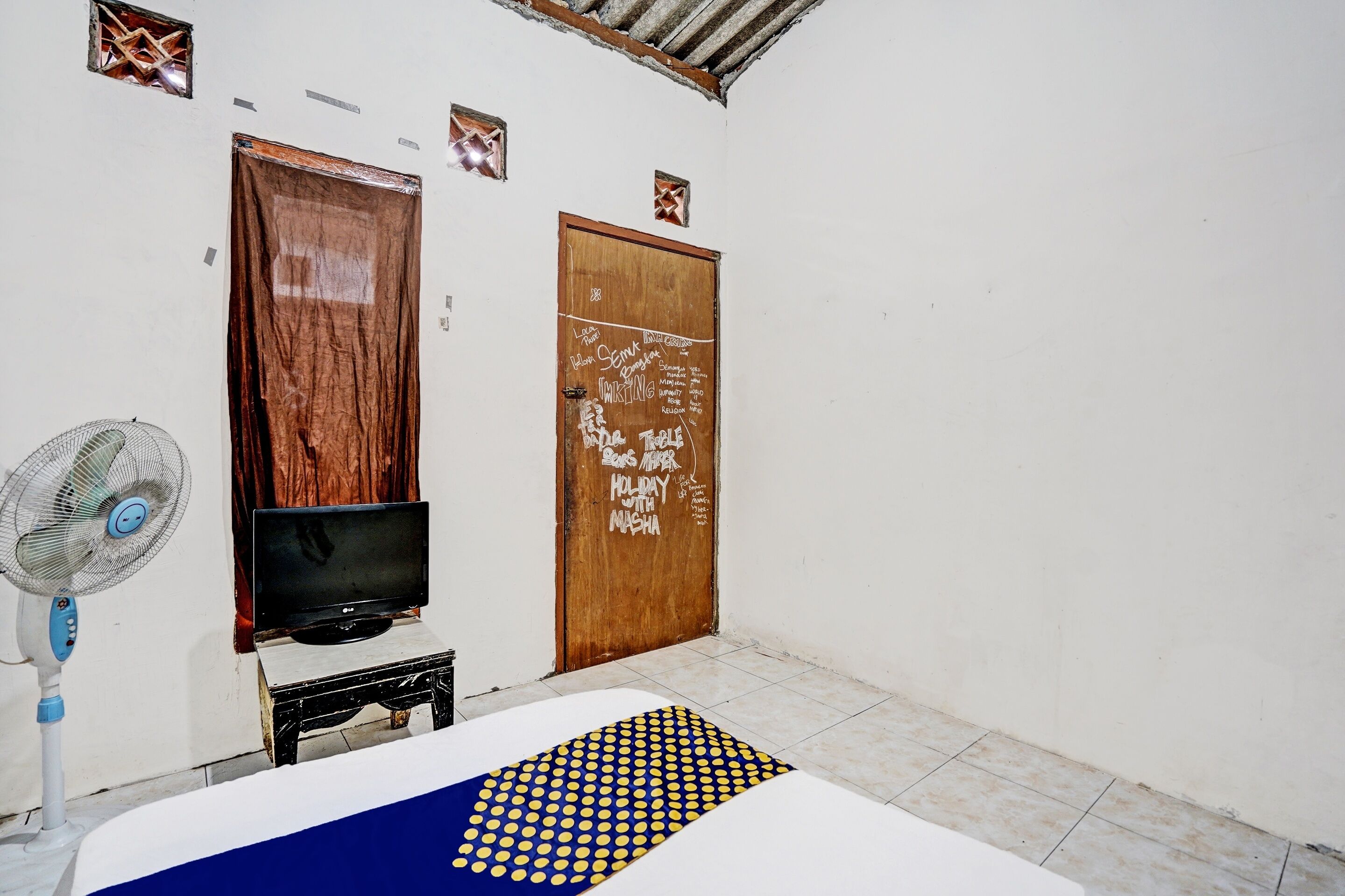 Una habitación con puerta de madera, un televisor sobre una mesita, un ventilador y una alfombra estampada.