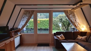 Une pièce chaleureuse avec un plancher en bois, un canapé, une table et un téléviseur. Des rideaux sont accrochés aux fenêtres, et on peut admirer la verdure à l’extérieur.