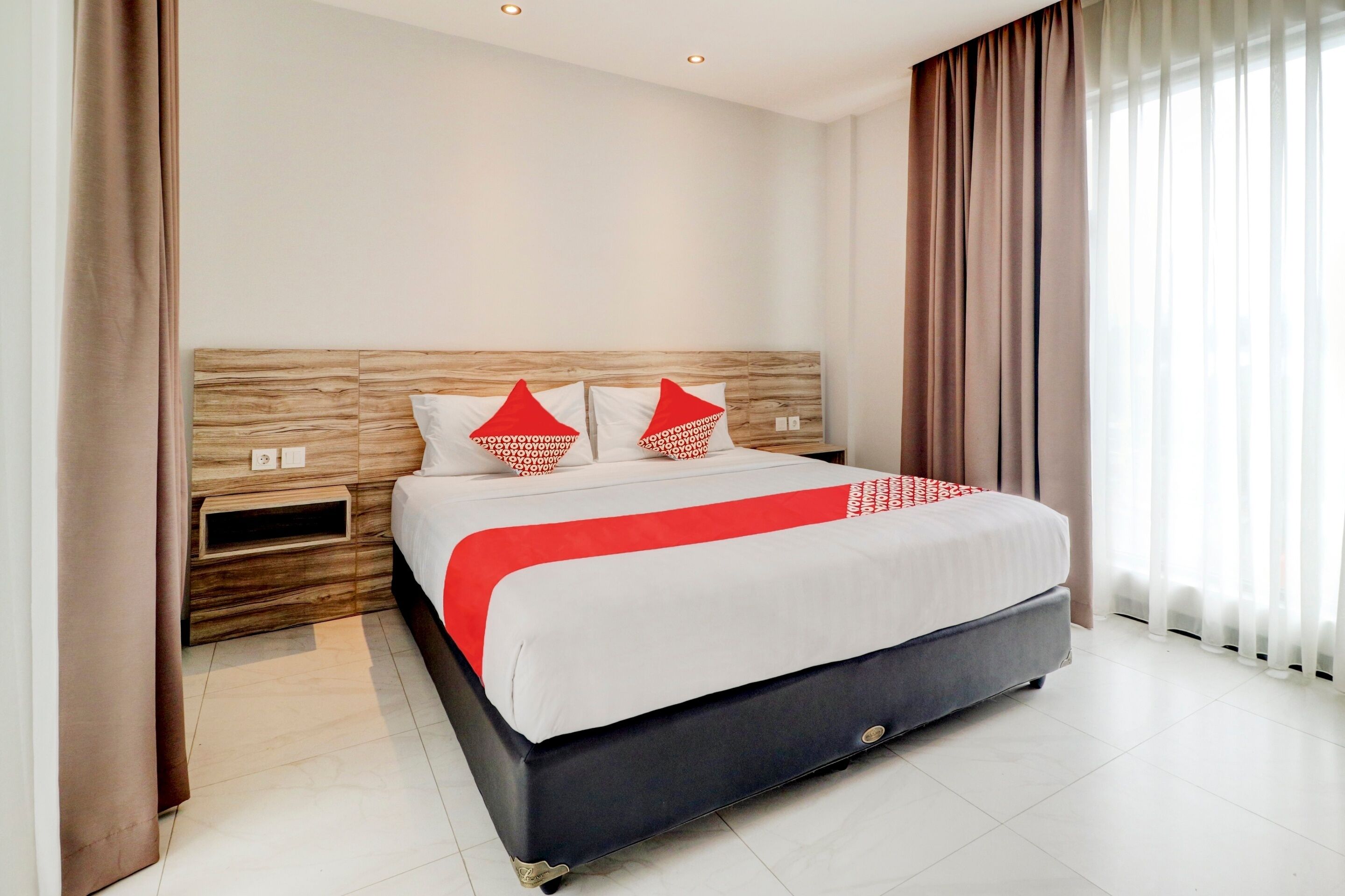 A modern hotel room with a wooden headboard, a large bed with a red and white bedspread, and a window with sheer curtains.