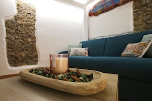 Una habitación con una mesa de madera que sostiene una vela y un cuenco de hierbas secas. Un sofá azul con cojines estampados se ve al fondo.