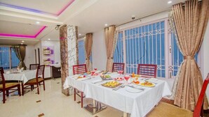 Un coin repas avec une longue table dressée pour un repas, entourée de chaises, avec une fenêtre et des rideaux en arrière-plan.