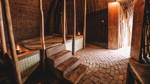 Ein rustikaler Raum mit Holzwänden und einem Strohdach. Treppen führen zu einer erhöhten Plattform mit einer Kerze.