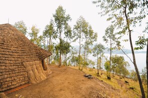 A rustic thatched-roof hut with a dirt path leading to a body of water, surrounded by trees and hills.