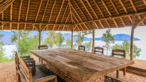 Ein überdachter Außenbereich zum Essen mit einem Holztisch und -stühlen, Backsteinböden und Blick auf ein Gewässer sowie Hügel im Hintergrund.