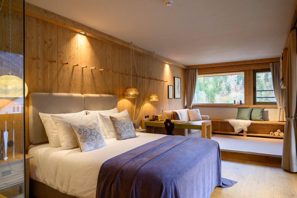 A bedroom with a large bed, a wooden wall, a window with a view of trees, and a bench by the window.