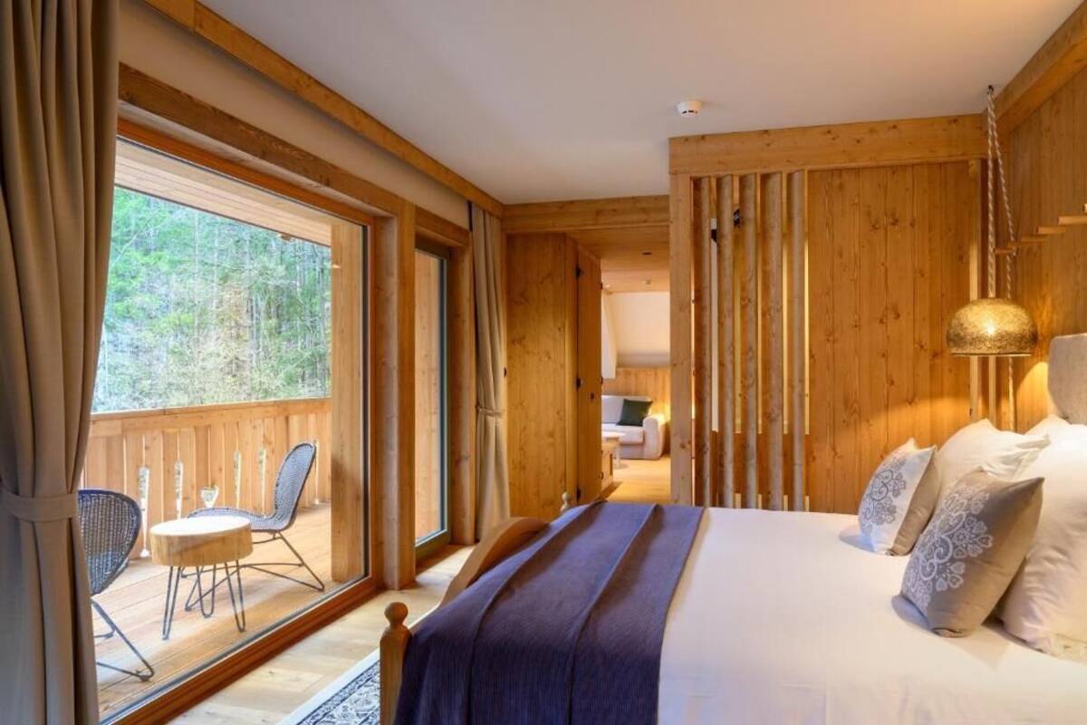 A wooden cabin bedroom with a large bed, a view of the forest through a window, and a balcony with chairs.