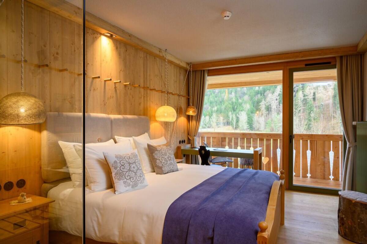 A wooden cabin bedroom with a large bed, a wooden desk, and a balcony with a view of the forest.