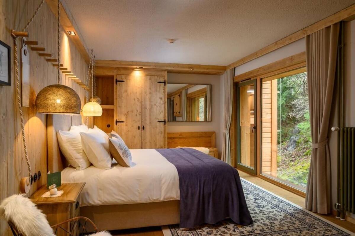 A bedroom with a large bed, wooden walls, a mirror, and a view of a forest through a sliding glass door.