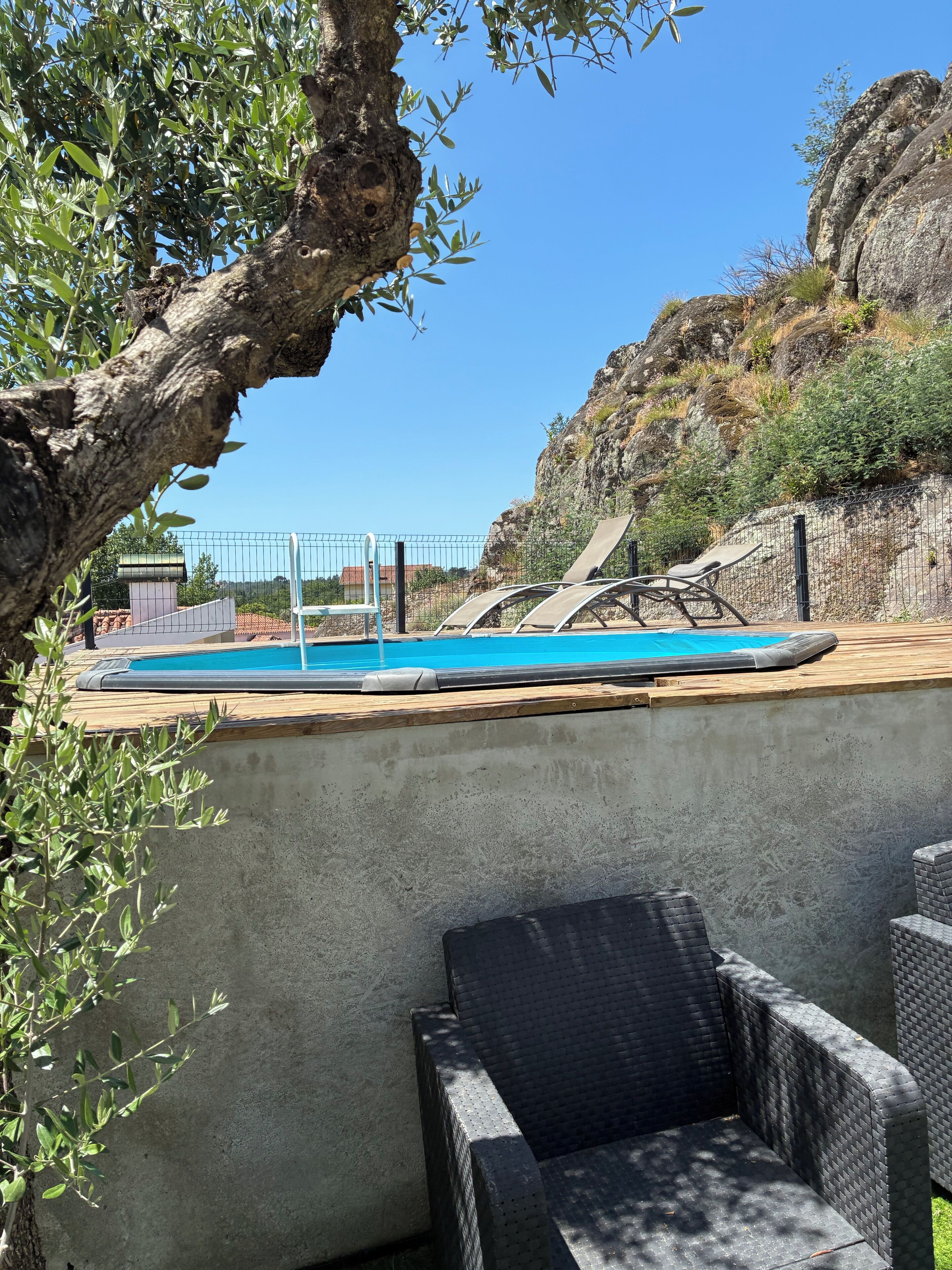 A swimming pool with a wooden deck, surrounded by a concrete wall, with outdoor furniture and a view of a rocky hillside.