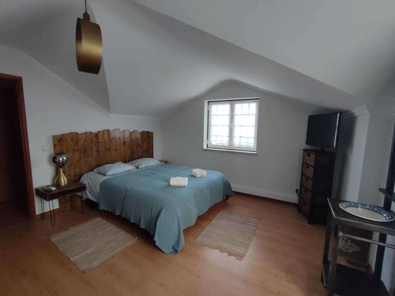 Une chambre avec un plafond en pente, un lit avec une literie bleue, une tête de lit en bois, une table de chevet avec une lampe, un téléviseur et une fenêtre avec des stores.