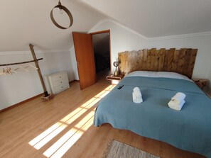 Quarto com cabeceira de madeira, cama com colcha azul, duas toalhas enroladas, mesa de cabeceira de madeira com abajur e guarda-roupa com cabides.