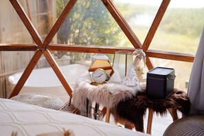 A cozy room with a wooden frame window, a bed with a white patterned bedspread, a fluffy white pillow, a small white lamp, and a black speaker on a furry stool.