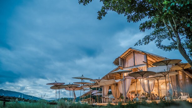Une construction rustique en bois, agrémentée de plusieurs parasols et rideaux, entourée de verdure et de collines.