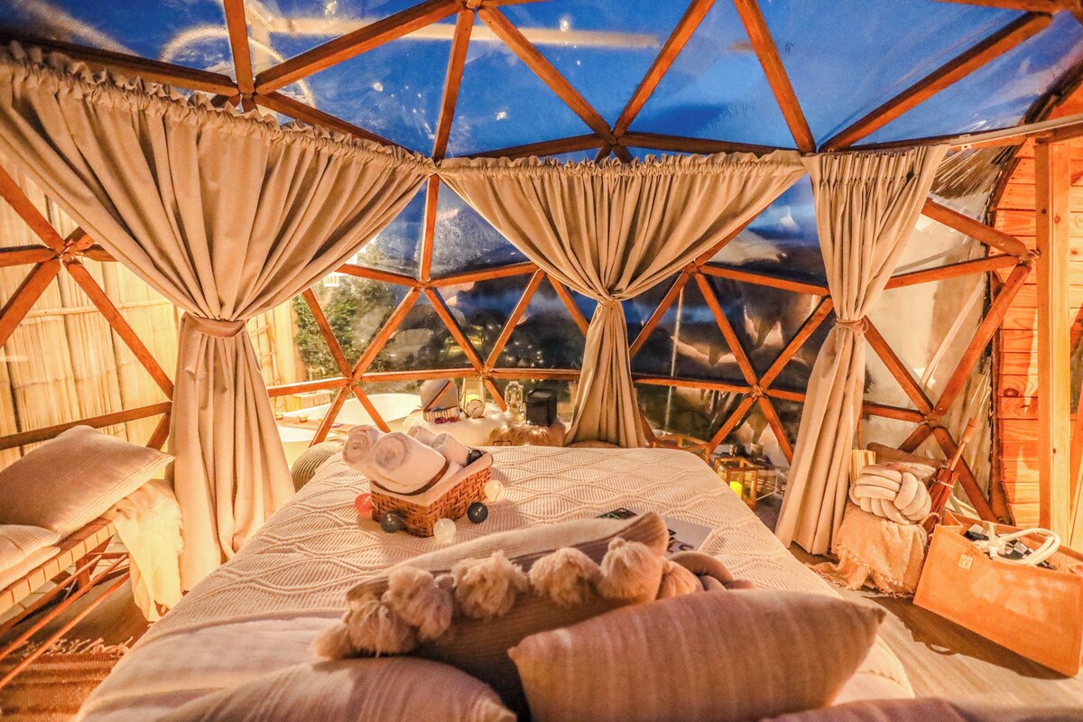 A cozy room with a bed, pillows, a small table with a candle, and a view through a geometric window structure.