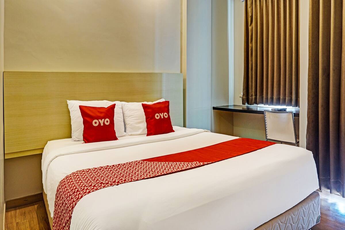 A hotel room with a neatly made bed, two pillows with 'OYO' branding, a red and white patterned throw, a bedside table with a lamp, and a window with curtains.