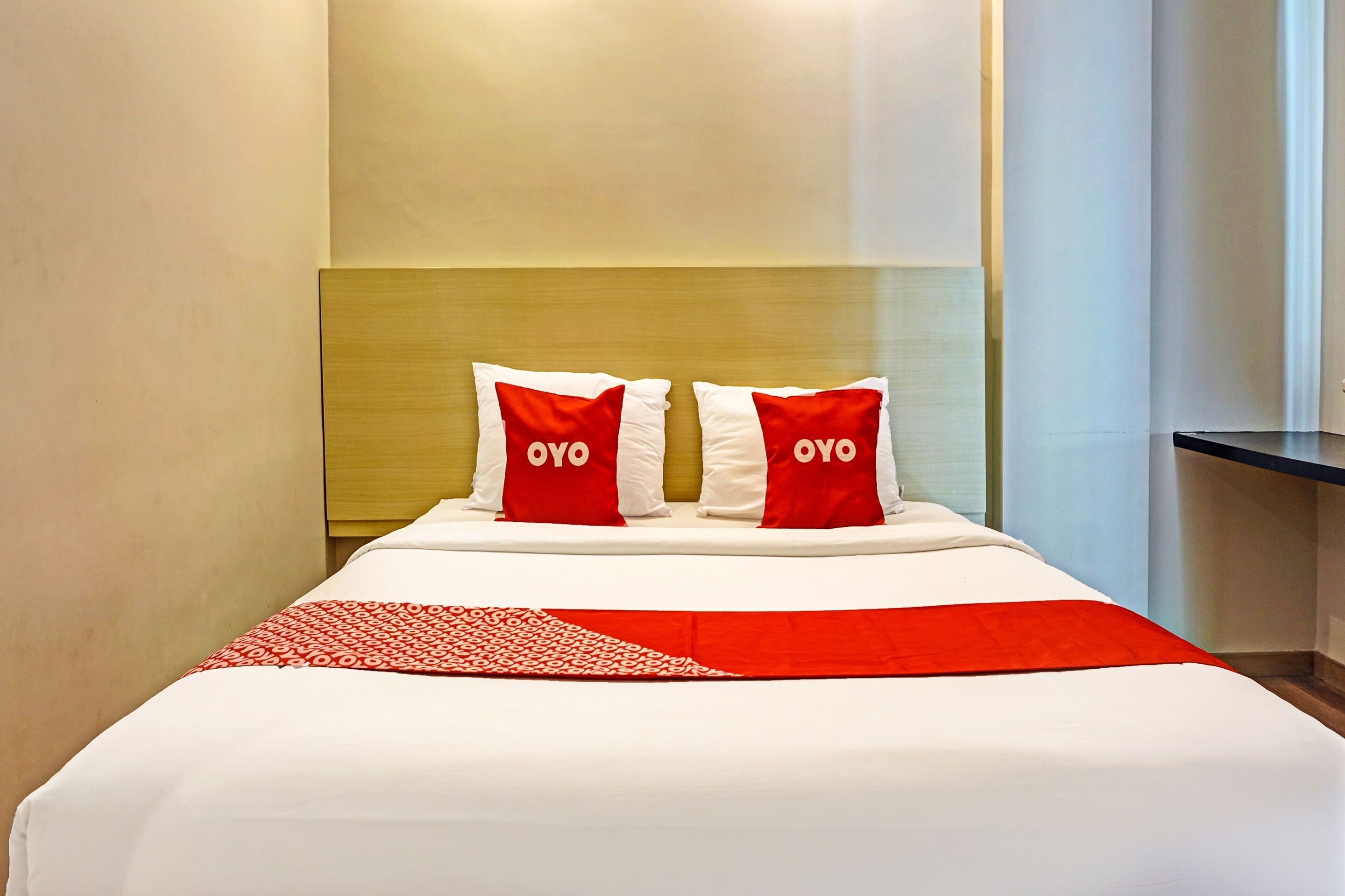 A hotel room with a bed, two pillows with "OYO" branding, a red and white patterned throw, and a wooden headboard.