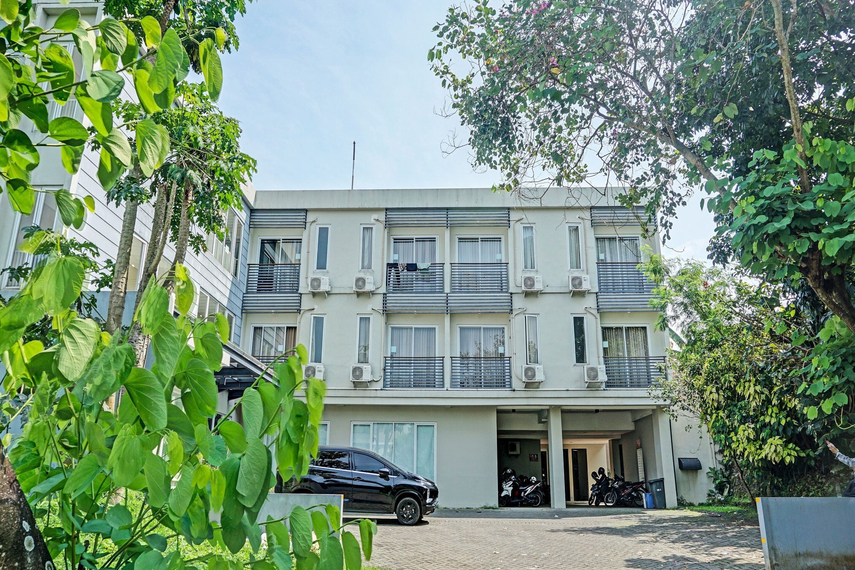 A multi-story residential building with balconies, air conditioning units, and a parking area with a black car and motorcycles.