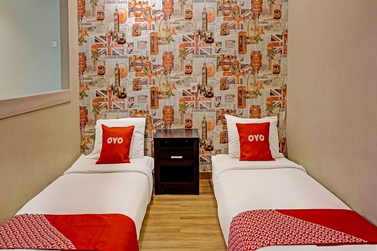 A hotel room with two beds, each with a red and white blanket and pillows with the word "OYO". The wall is decorated with a travel-themed wallpaper featuring London landmarks.