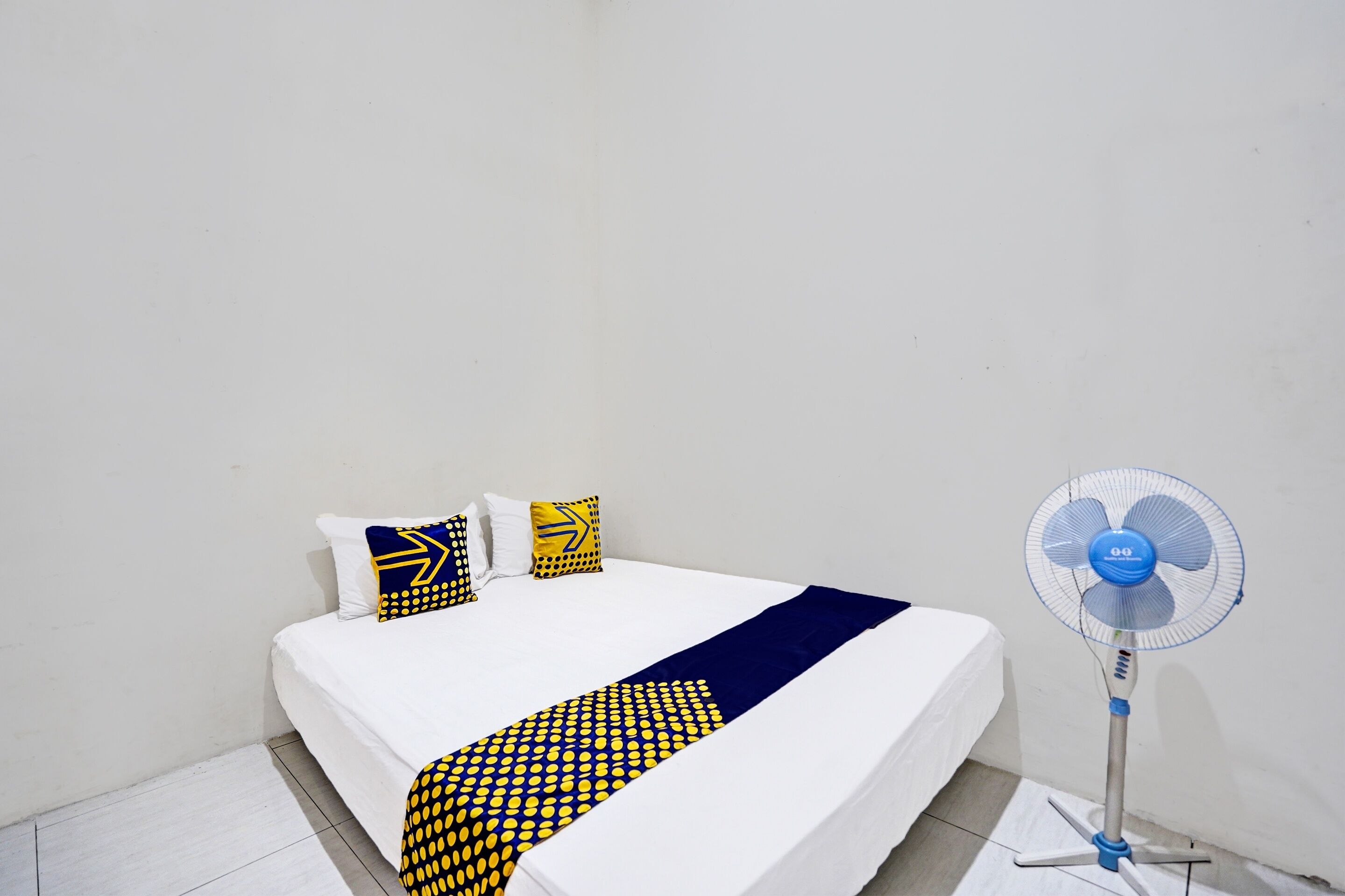 A bed with a white mattress, a blue and yellow patterned blanket, and three pillows with geometric designs. A standing fan is positioned to the right of the bed.