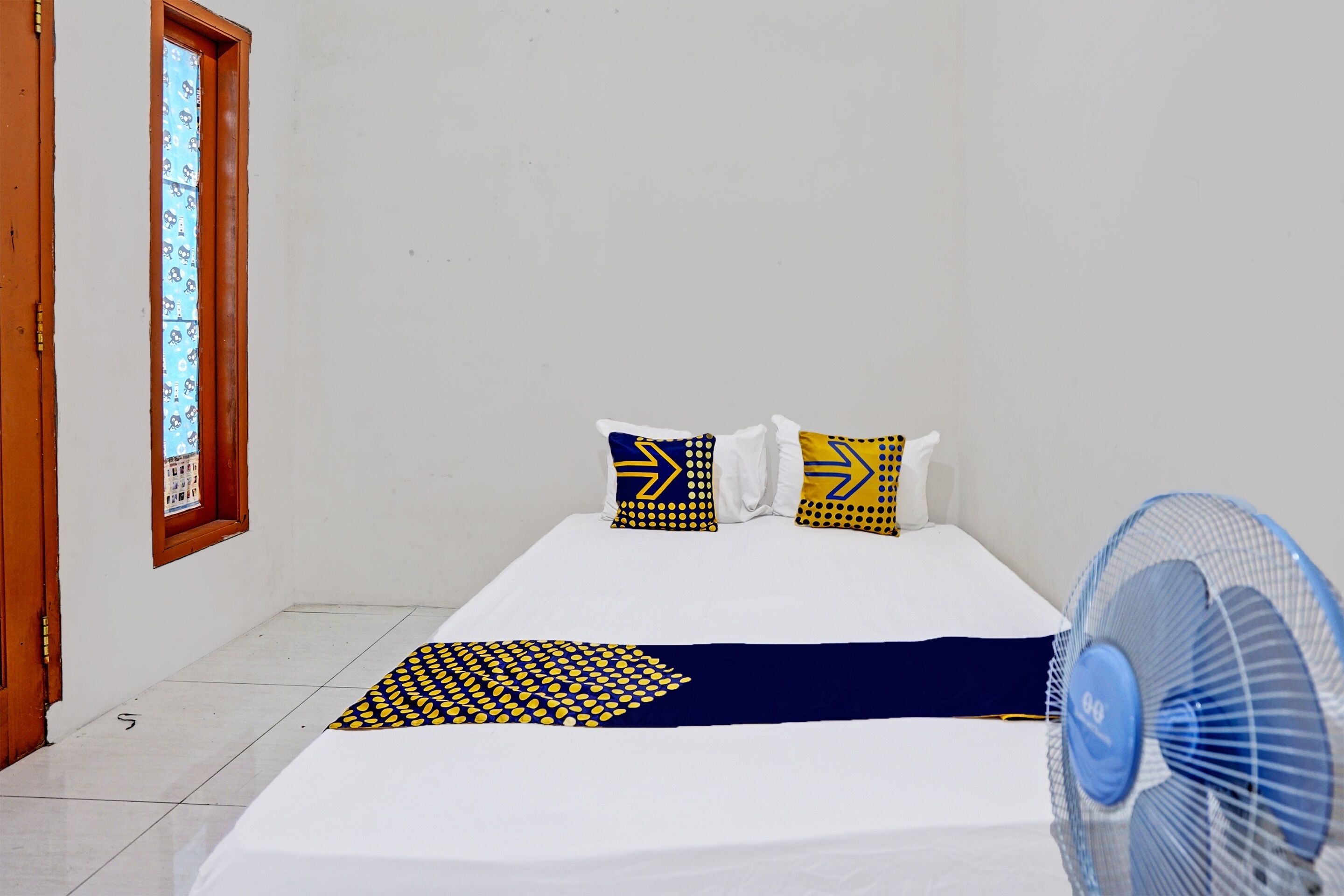 A single bed with a white mattress, two pillows with yellow and blue patterns, a blue fan, and a window with a patterned view.