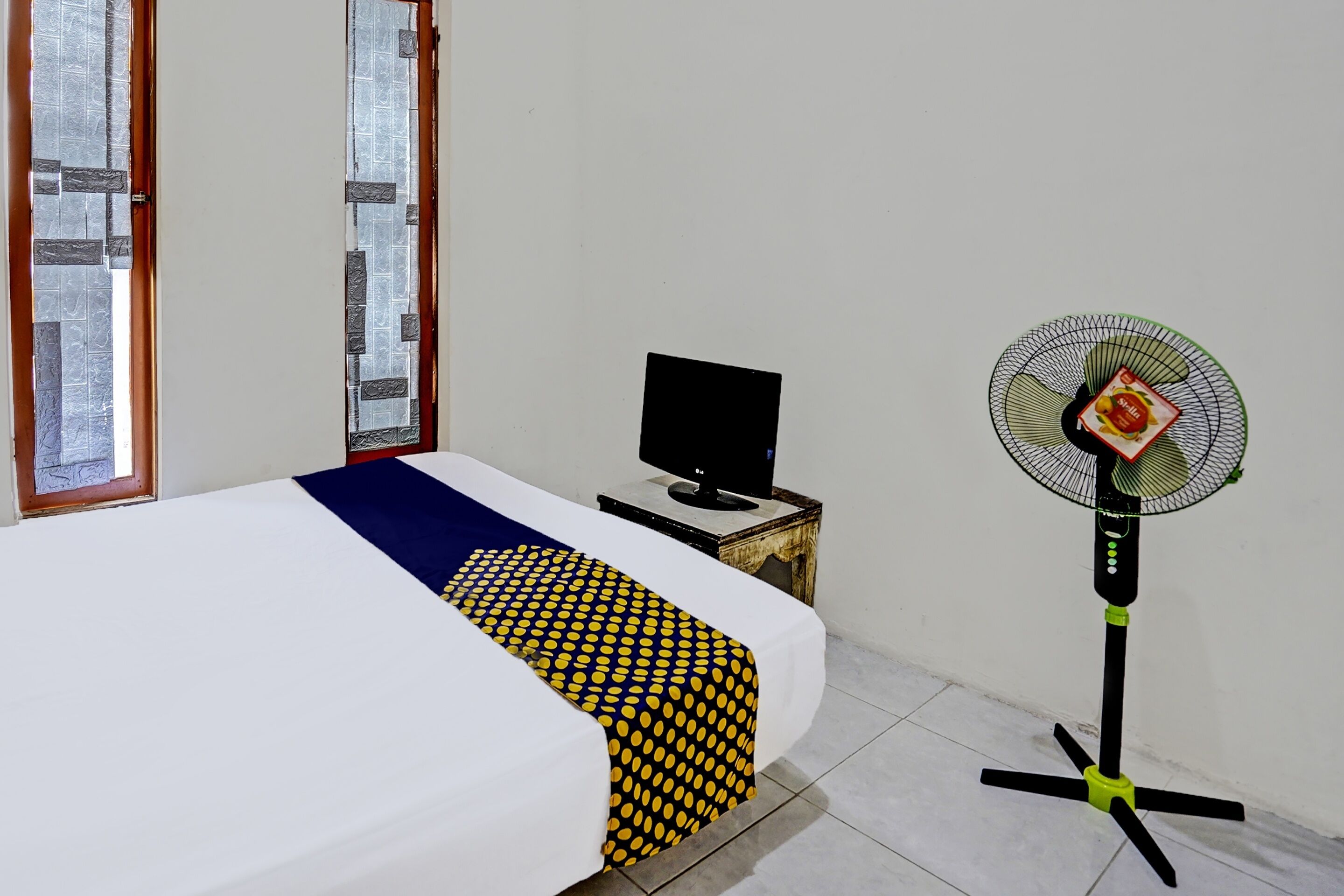 Een slaapkamer met een bed, een televisie op een nachtkastje en een staande ventilator.