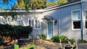 Une maison de plain-pied avec un porche à l’avant, une porte bleue et un petit jardin agrémenté de plantes et d’une chaise en métal.