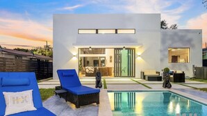 A modern house with a swimming pool, blue lounge chairs, and a well-lit interior.