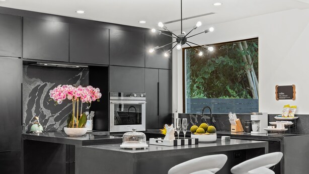 A modern kitchen with dark cabinetry, a marble backsplash, a central island with bar stools, and a large window with a view of greenery.