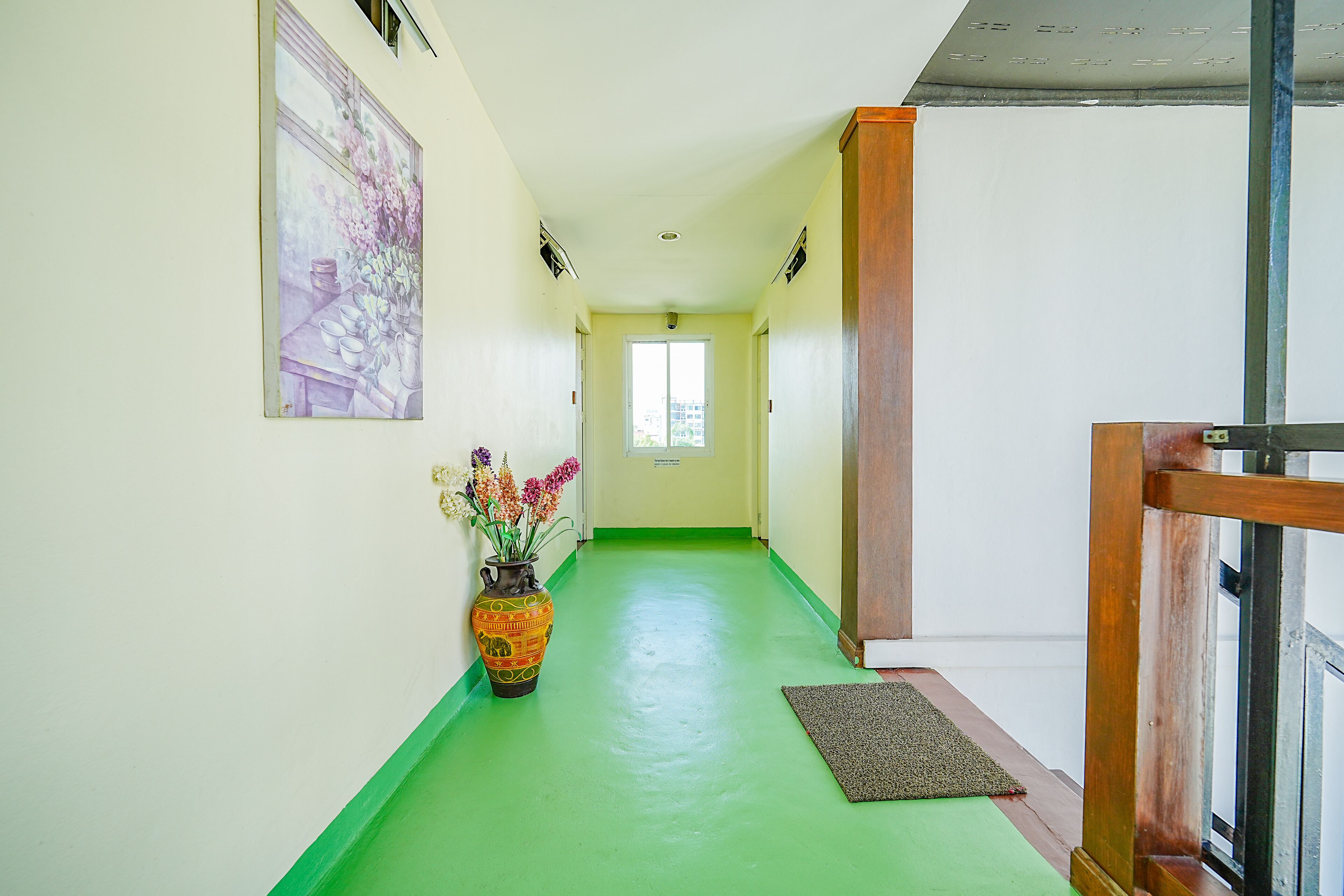 A hallway with a green floor, a wooden railing on the right, and a vase with flowers on the left.