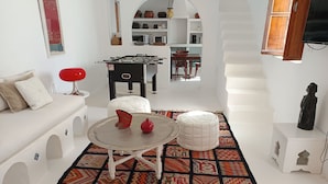 Una sala moderna con un sofá, un reposapiés y una mesa redonda con una granada. Hay una alfombra estampada en el suelo y una zona de cocina al fondo.