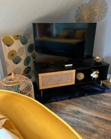 Uma sala de estar moderna com uma TV de tela plana sobre um suporte preto, uma parede decorativa com padrões de folhas, uma mesinha lateral trançada e um tapete felpudo azul.