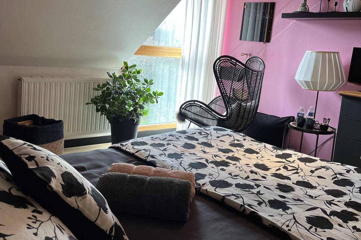 A bedroom with a bed, a chair, a table, a lamp, a plant, and a window with curtains.