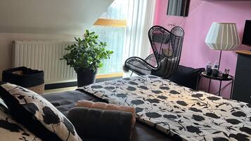 Ein Schlafzimmer mit Bett, Stuhl, Tisch, Lampe, Pflanze und einem Fenster mit Vorhängen.